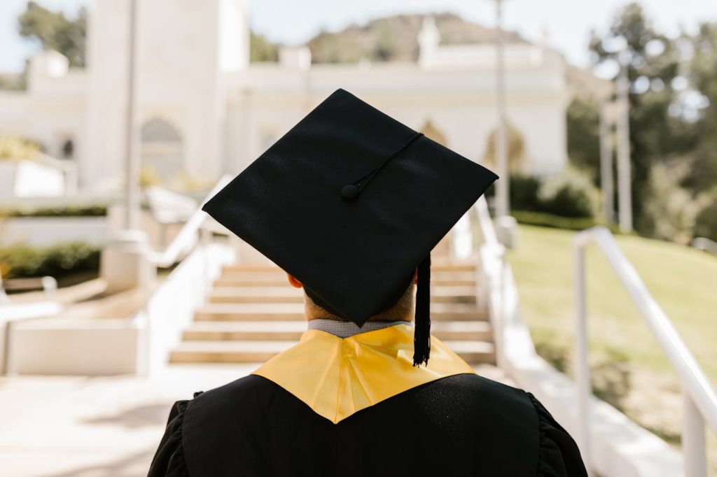 a-graduate-wearing-a-mortarboard-and-a-graduation-gown-7713511 Back view of a graduate in a cap and gown standing outdoors facing a building.