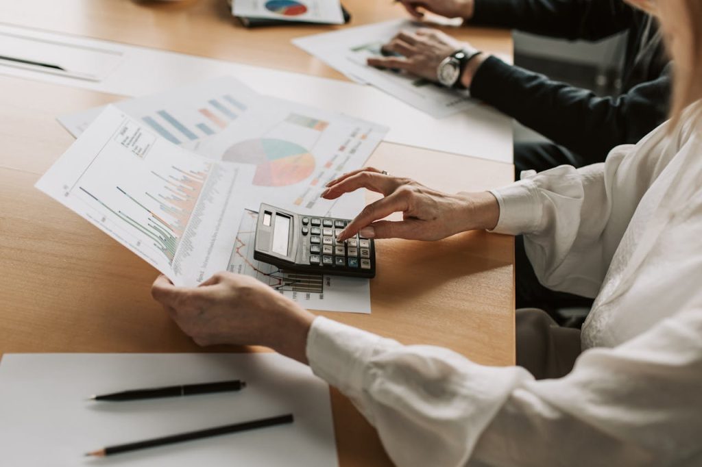 a-person-using-a-calculator-7433832 Hands performing financial calculations with charts and a calculator at a meeting table.